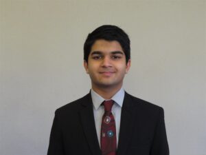 2021-22 Cutler-Bell Student Winner Harshal Bharatia is a young man in a suit and red tie. He has short black hair, medium brown skin and is smiling against a plain backdrop.