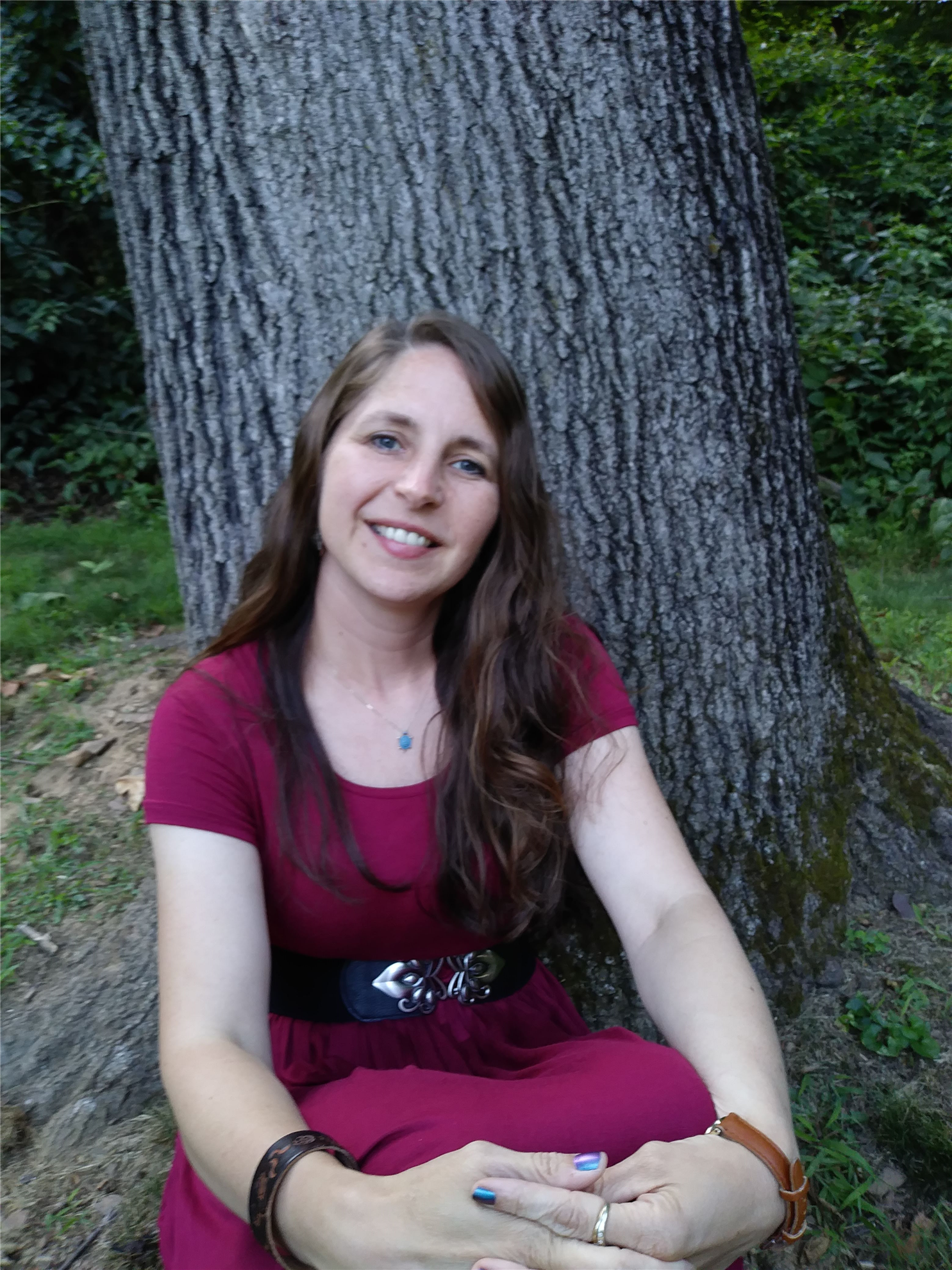 Jacqueline Corricelli headshot, a white woman with a maroon dress, long brown hair, sitting in front of a tree