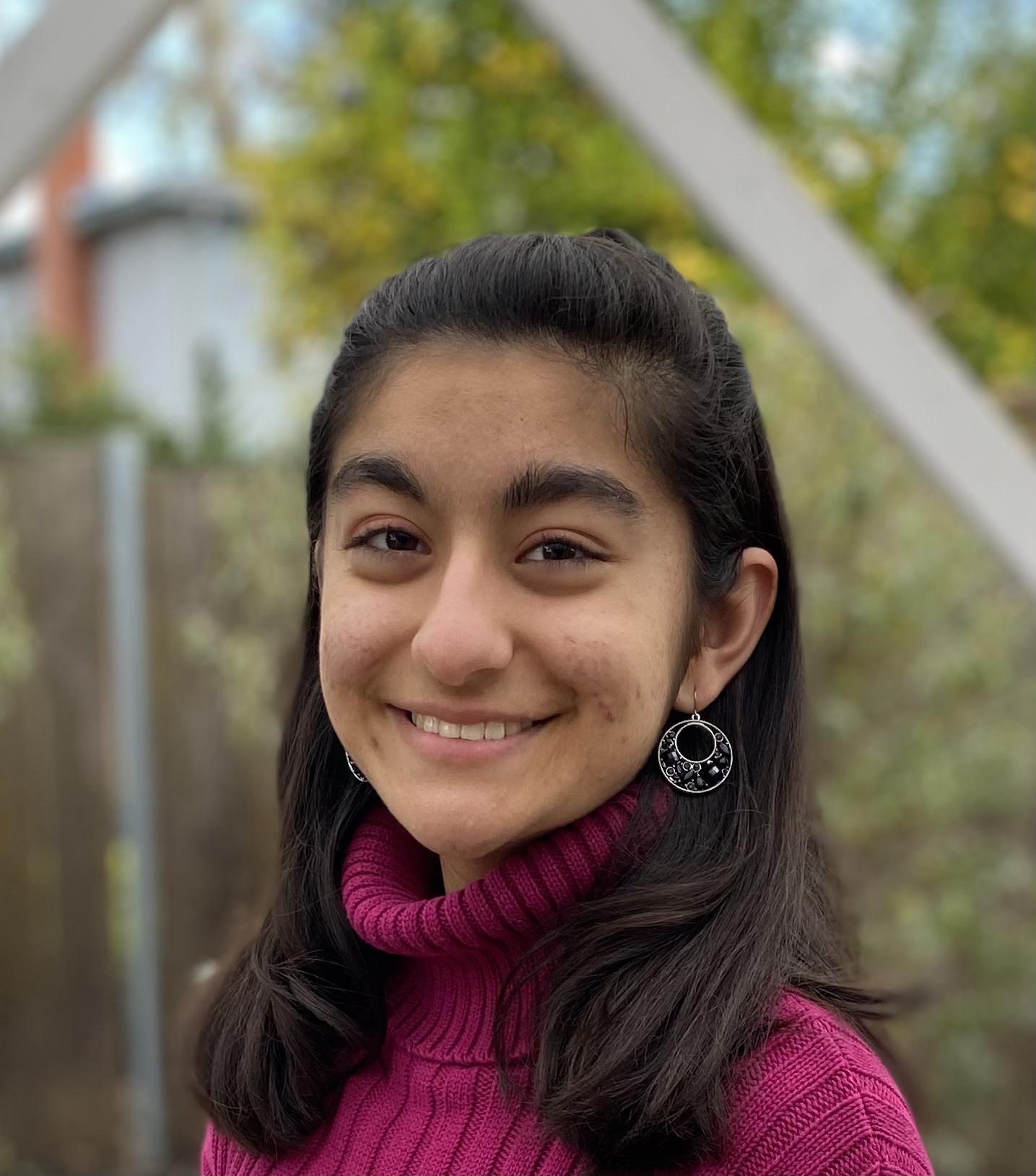 2021-22 Cutler-Bell Student Winner Hiya Shah is a young woman with long, straight black hair in a half-up hairstyle. She has medium brown skin and brown eyes. She wears a pink turtleneck sweater, silver hoop earrings and is smiling at the camera.