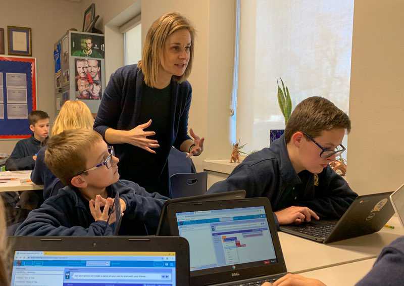 Rebecca Luebker is between two young boys with laptops in front of them and appears to be amid an explanation during a CS class