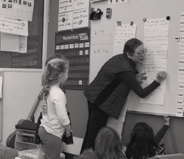 A teacher writes something on paper that is on a whiteboard while a kindergarten class looks on