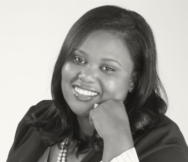 Black and white headshot of Leontae Gray Ward, a black woman with straight black hair, black blazer, and pearl necklace. She is smiling at the camera and posing with her face gently resting on her hand.
