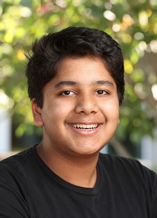 2021-22 Cutler-Bell Student Winner Yash Narayan is a young man with brown eyes, short black hair and medium brown skin. He wears a black t-shirt and is smiling. The background makes it clear he is outside in the sun.
