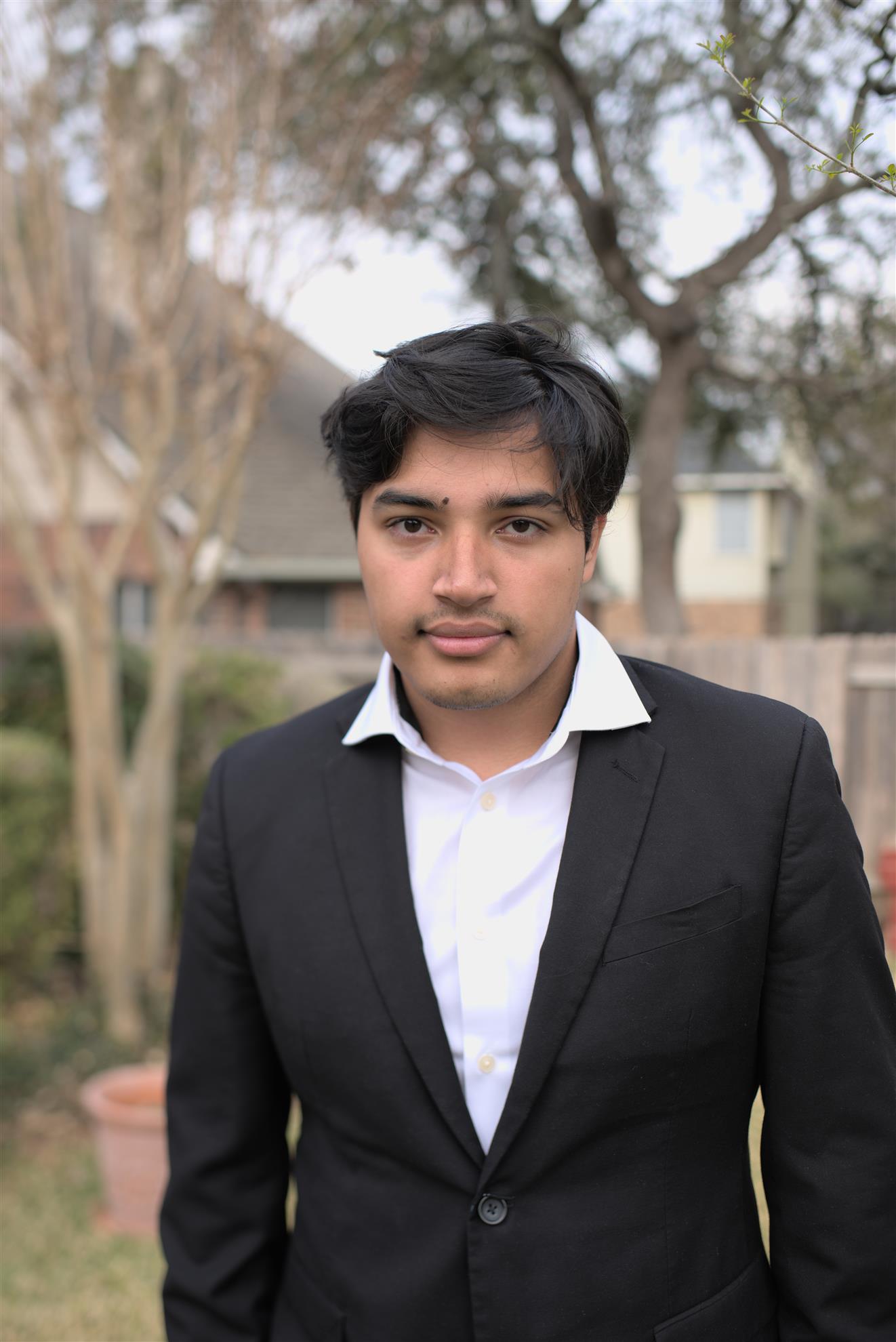 2021-22 Cutler-Bell Student Winner Shoumik Roychowdhury is a young man with a black suit jacket and white button down shirt. He has medium brown skin, short black hair and brown eyes. He stands outside and looks into the camera with hands in his pockets.