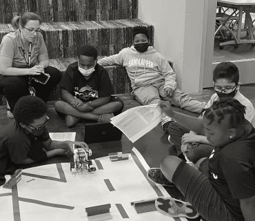 Black and white photo of a computer science teacher with a group of 5 middle school boys designing a course in robotics. They have just set their robot down on the course.