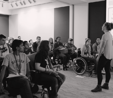 a room full of people with a woman at the front facing and talking to them. There is a wheelchair user in the front row.