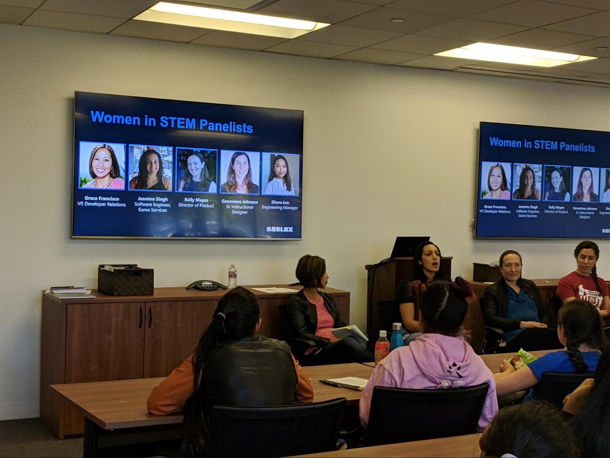 Laura Ramirez at a meeting with her school's teacher-led girls group. A TV screen reads "women in STEM panelists" behind the group