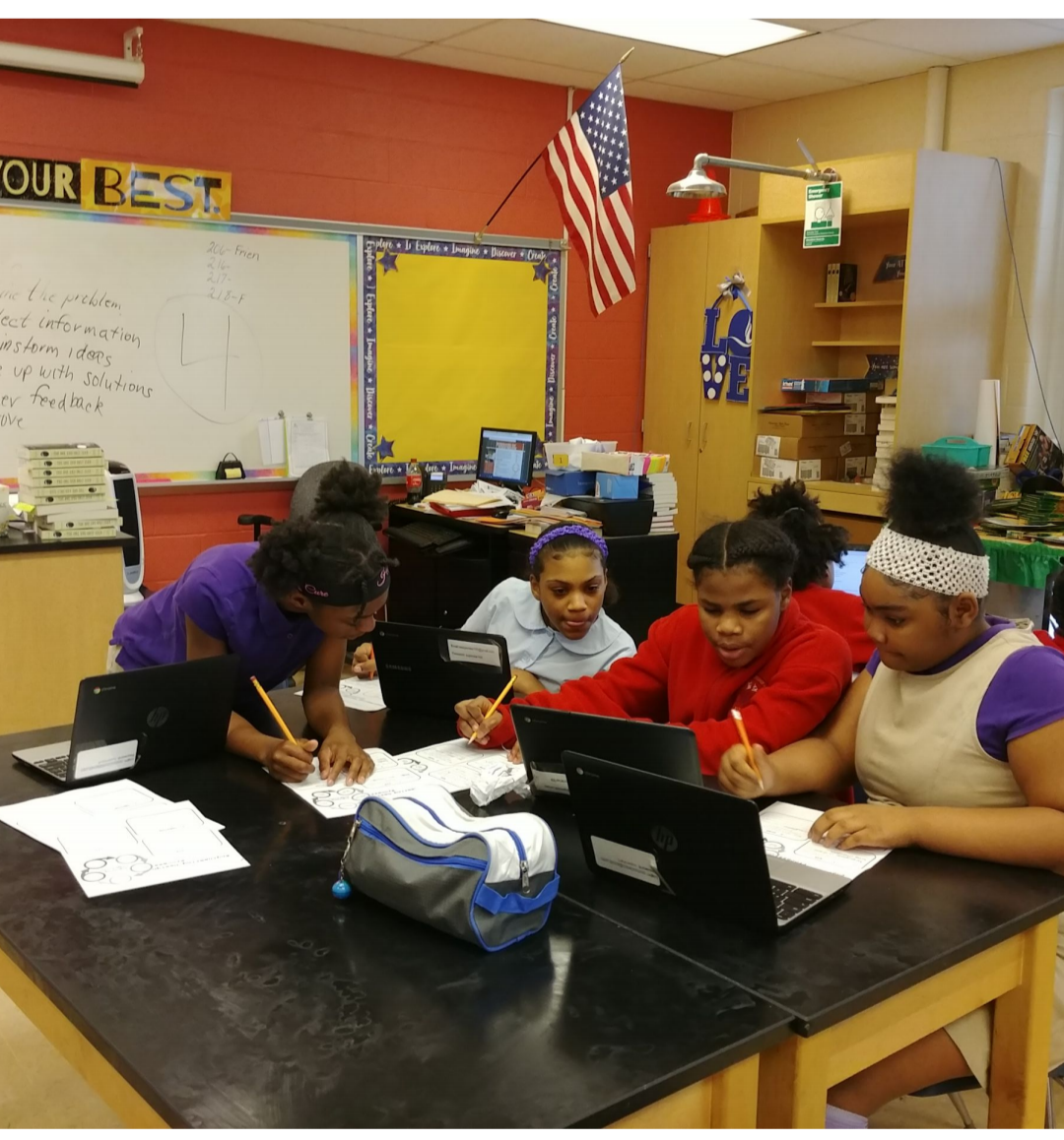 A table with four African American girls in computer science class, all writing something down while looking excitedly at their laptops