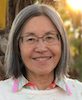 Headshot of Jane Prey, an older woman with eastern Asian features. She has straight silver hair cut into a bob, medium-light skin tone, and is wearing rectangular glasses and a white sweater. She is smiling to the camera with the sun behind her.