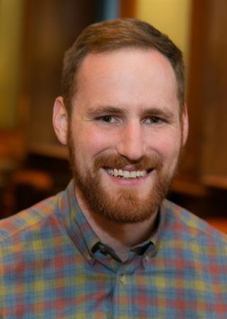 Headshot of Jake Baskin, next executive director of CSTA. He is a caucasian mane with short ginger hair and a close cropped ginger beard. He is wearing a red, blue and yellow plaid button down shirt and is smiling into the camera.