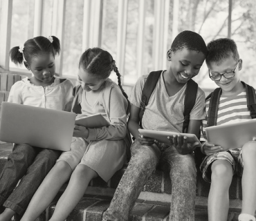 Four kids with backpacks on, all smiling and interacting with a tablet or computer screen.