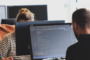 Two people sit in front of computers coding. 