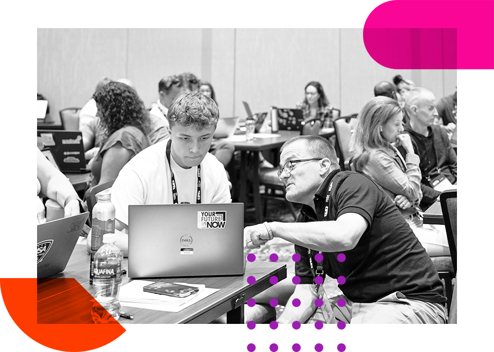 A man leans overs and helps someone on a computer during a conference session