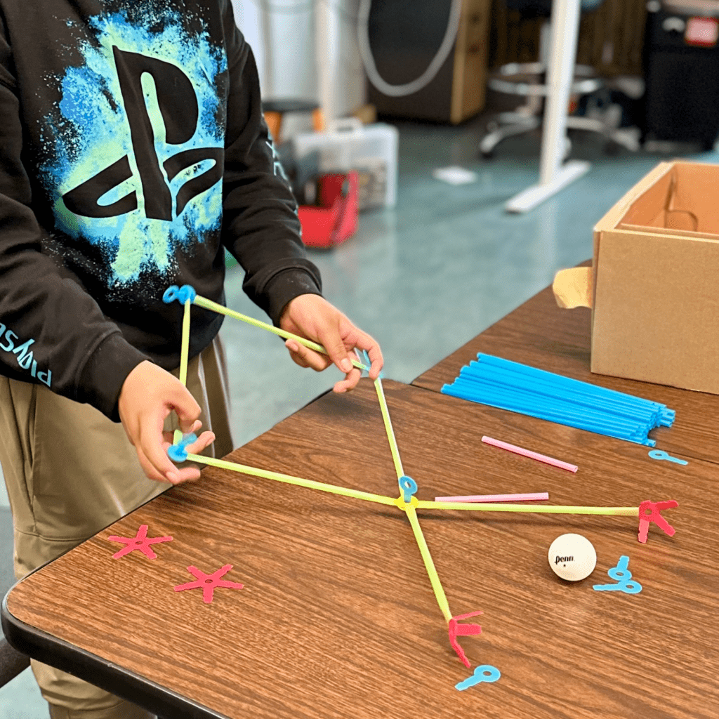 A student holding the beginnings of a straw construction on a table crowded with straw connectors and half-built structures.